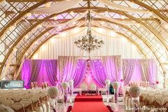 A mandap in a grand ballroom with pink lighting, white drapes, and gold accents.